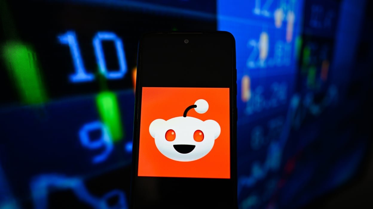 a Reddit logo is seen displayed on a smartphone with stock market percentages on the background