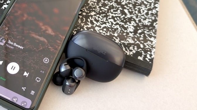 black shokz opendots one earbuds propped against iphone and composition notebook